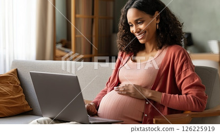 Happy pregnant black mom working remotely on laptop. Online video call. The smiling mom embodies the spirit of working mothers as she connects online during her pregnancy. Happy pregnant black mom working remotely on laptop. Online video call. The smiling mom embodies the spirit of working mothers as she connects online during her pregnancy. 126225919
