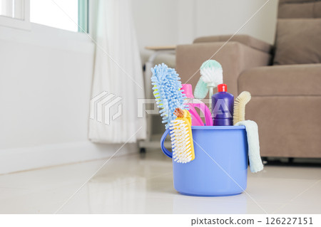 Cleaning essentials at home. A blue bucket containing detergent bottles, brushes, and other supplies sits on the floor in the living room, helping with routine housework and general housekeeping. 126227151