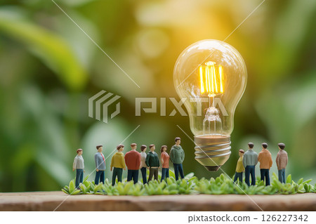 A group of small figurines surrounds a glowing light bulb on green grass, evoking themes of innovation, teamwork, and sustainability in a creative and conceptual composition. Ideas and unity concept 126227342