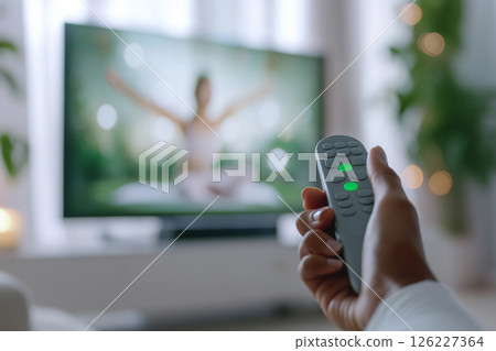 A close-up of a hand holding a remote control, focusing on a yoga instructor displayed on a TV in a modern and serene home setting, emphasizing fitness and relaxation. Blurred background 126227364