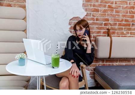 Businesswoman talking on phone at cafe with laptop and green smoothie Businesswoman talking on phone at cafe with laptop and green smoothie 126230558