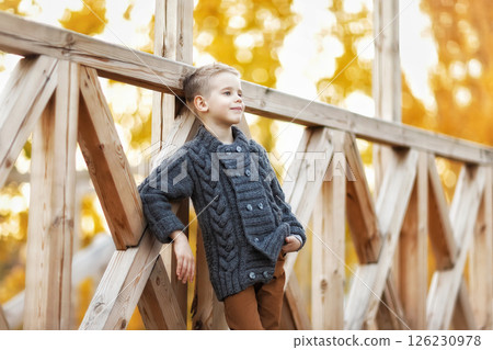 Little smiling caucasian child wearing jacket and trousers is posing at wooden bridge outdoors background. 126230978