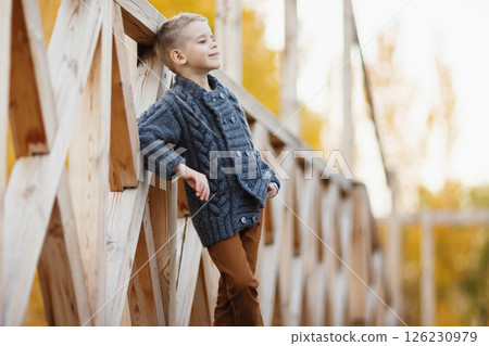 Little happy caucasian child wearing jacket and trousers is posing at wooden bridge outdoors background. Little happy caucasian child wearing jacket and trousers is posing at wooden bridge outdoors background. 126230979