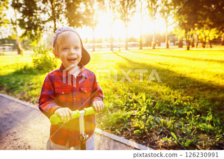 Happy little boy is riding scooter and smiling at summer sunny park background. Happy little boy is riding scooter and smiling at summer sunny park background. 126230991