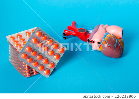 Stack of red pills against the background of a heart model and a blue background. Concept of treating diseases in cardiology, angina. Vitamins for the heart. Pills for arrhythmia. Copy space for text 126232900