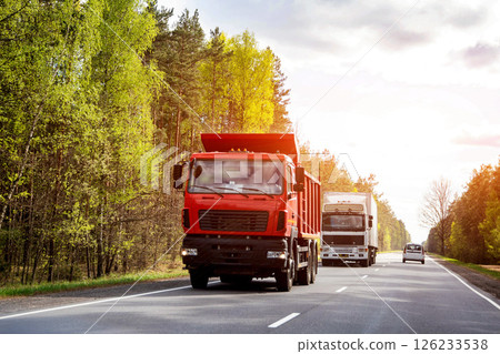 Red dump truck in a convoy of trucks transports cargo along a country road in summer against the backdrop of the sun. Copy space for text 126233538