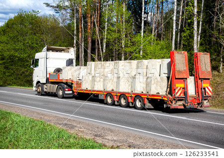 A white truck with a lowboy semitrailer transports concrete products along a country road against a forest background. Oversized cargo. Copy space for text A white truck with a lowboy semitrailer transports concrete products along a country road against a forest background. Oversized cargo. Copy space for text 126233541