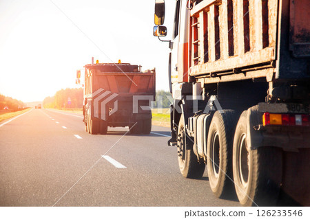 Orange road dump trucks drive along the highway after delivering asphalt against the backdrop of sunset in summer, road repair. Copy space for text, industry 126233546