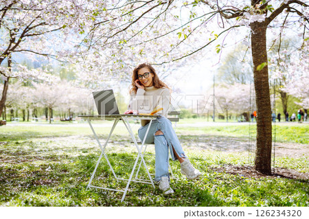 A young woman at a table in a blooming park works with a laptop. Freelancing, weekend concept. 126234320