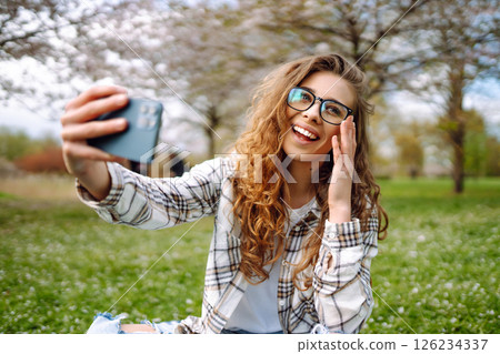 Young woman takes selfie against background of flowering trees in park. Concept of relaxation, fun. 126234337