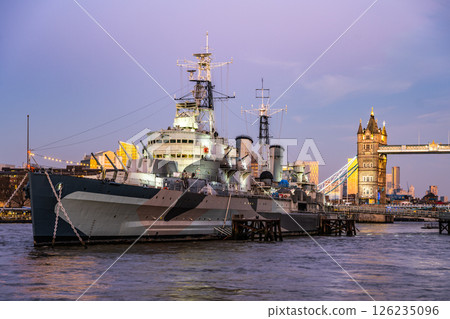 HMS Belfast is anchored on the Thames River in London during the evening. The warm glow from the city highlights the iconic ship and the nearby Tower Bridge, creating a picturesque view. 126235096
