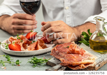 Hands holding a glass of wine and a wooden board with different kinds of cheese and ham, prosciutto, jamon salami, Antipasto Dinner or aperitivo party 126235758