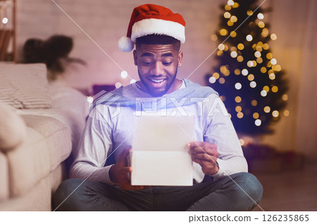 Christmas gift. Happy black guy opening box, sitting against Christmas tree and wearing santa hat 126235865
