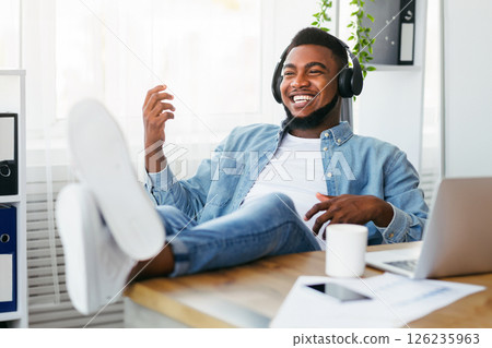 Break at work. Cheerful african american employee listening music in headphones and playing virtual guitar, relaxing at workplace 126235963