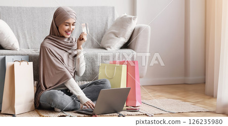 Online Shopping Concept. Young arab girl in hijab typing credit card data on laptop, purchasing modest clothes, sit among bright shopper bags on floor Online Shopping Concept. Young arab girl in hijab typing credit card data on laptop, purchasing modest clothes, sit among bright shopper bags on floor 126235980