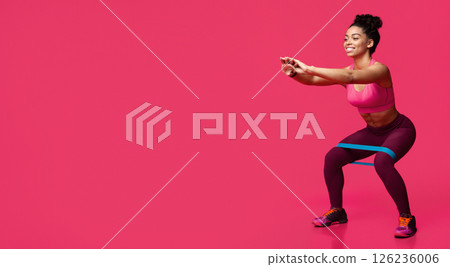Young sporty smiling black woman doing stretching work out with elastic bands, squating over red studio background 126236006