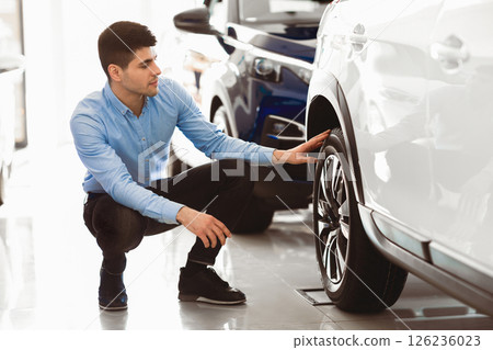Examining Car. Handsome man sitting near brand new auto and checking wheel rims and tire, copy space Examining Car. Handsome man sitting near brand new auto and checking wheel rims and tire, copy space 126236023