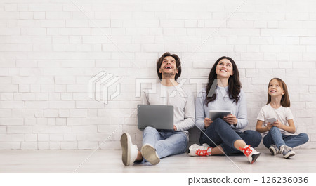 Great Online Bonus For All Family. Father, Mother And Daughter Sitting On Floor With Diverse Gadgets In Hands And Looking Upwards, Free Space 126236036