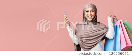 Cashback concept. Portrait of exited arabic woman in hijab holding smarphone and bright paper shopping bags and looking at camera, pink background Cashback concept. Portrait of exited arabic woman in hijab holding smarphone and bright paper shopping bags and looking at camera, pink background 126236051