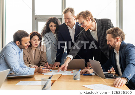 A group of business professionals are gathered around a table in a modern office, collaborating on a project. They are reviewing documents and using laptops. A group of business professionals are gathered around a table in a modern office, collaborating on a project. They are reviewing documents and using laptops. 126236100
