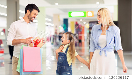 A father holding shopping bags and a gift with a red bow reveals it to his excited daughter, as his wife walks beside them. The three are shopping in a modern mall A father holding shopping bags and a gift with a red bow reveals it to his excited daughter, as his wife walks beside them. The three are shopping in a modern mall 126236117