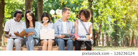 Multiethnic friends studying for exams together outdoors, sitting on bench in campus, having fun together 126236124