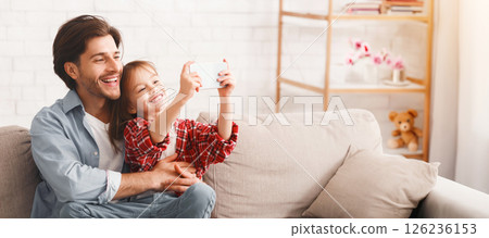 Preschool girl taking selfie with her cheerful father, sitting on sofa at home, copy space 126236153