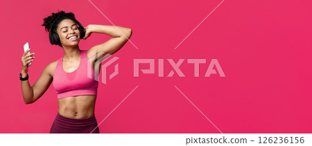 Happy african fitness girl enjoying music during exercising, holding phone and wearing wireless earphones over red background, awesome sport music application concept Happy african fitness girl enjoying music during exercising, holding phone and wearing wireless earphones over red background, awesome sport music application concept 126236156