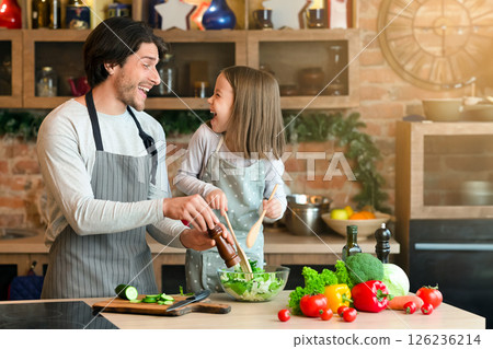 Happy father and little daughter cooking vegetable salad in kitchen, adding seasoning and laughing together while preparing lunch at home 126236214