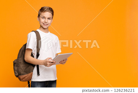 Education and modern technologies. Schoolboy with backpack using tablet computer and smiling on orange background, empty space 126236221