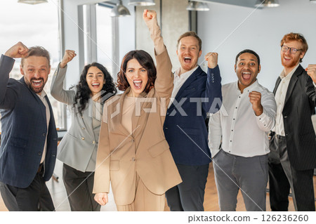 A diverse group of six business professionals, dressed in professional attire, stand together in a modern office setting. They are all smiling and raising their fists in the air 126236260