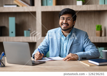 A smiling Indian man with a beard and glasses sits at a desk in a modern office, working on a notebook while looking directly at the camera. He is wearing a blue suit jacket and a light blue shirt. 126236281