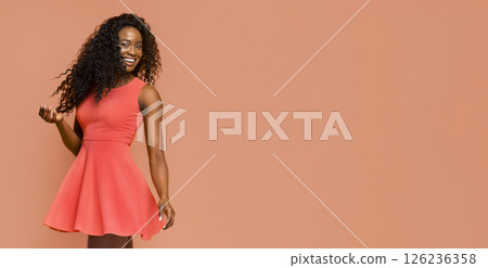 African American woman in a coral dress standing in front of a peach background. She has long, curly black hair and is smiling. Her arms are slightly raised as she looks towards her left side. 126236358