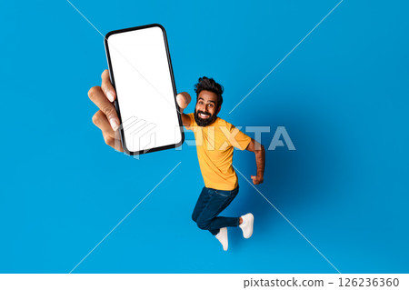 Indian man with a beard is jumping in the air, smiling, and holding a smartphone in front of him with a blank screen. He is wearing a yellow t-shirt and jeans against a bright blue background. 126236360