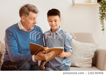 Elderly Grandfather And Grandson Boy Reading Book Sitting Together On Sofa At Home. Family Weekend. Selective Focus Elderly Grandfather And Grandson Boy Reading Book Sitting Together On Sofa At Home. Family Weekend. Selective Focus 126236406