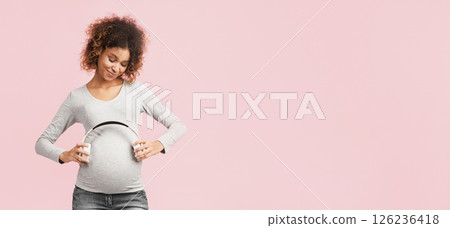 Pregnancy and music. Afro expectant woman holding earphones near belly over pink studio background 126236418