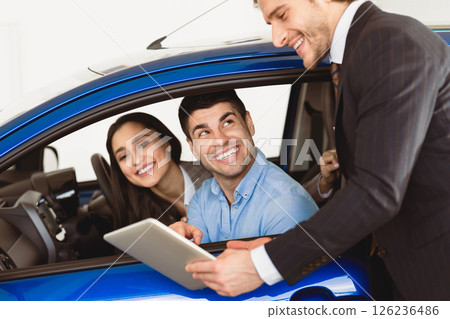 Check Out The Characteristics. Happy couple sitting in salon of new car, vendor showing tablet to them 126236486