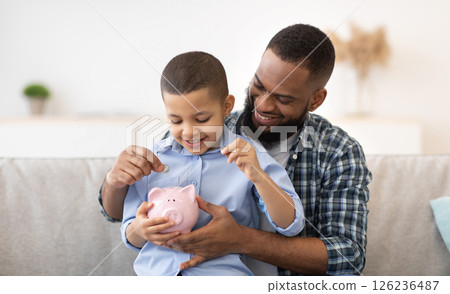 Financial Literacy. Cheerful African Father And Son Putting Money In Piggybank Sitting On Sofa At Home. Daddy Teaching His Child Budget Planning, Keeping Personal Savings Safety 126236487