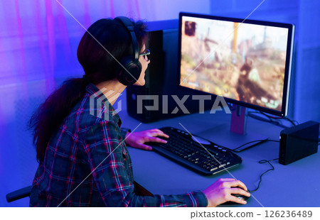 A young woman sits in a chair in front of a computer, wearing headphones and playing a video game in a room lit in blue light. Her hands are on the keyboard and mouse. 126236489
