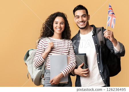 Study Abroad. Cheerful Young Arab Man And Woman With Backpacks Holding British Flag, Portrait Of Happy Smiling Students With Union Jack And Workbooks Over Yellow Studio Background, Copy Space 126236495