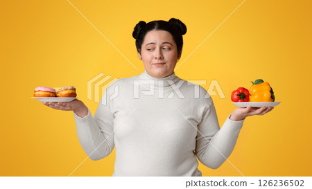Healthy Vs Unhealthy Food. Young obese girl choosing between fresh vegetables and donuts, keeping hands as a scales over yellow background, copy space 126236502