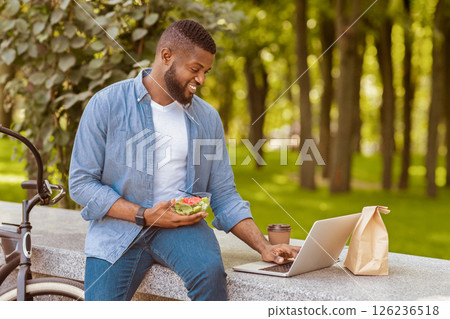 Joyful african american guy using laptop and eating lunch outdoors, sitting on bench in park with his bicycle parked by, free space 126236518