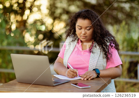 A young woman sits at an outdoor table, focused on writing notes while using a laptop. She enjoys the fresh air and natural surroundings, wearing a pink shirt and a plaid vest. A young woman sits at an outdoor table, focused on writing notes while using a laptop. She enjoys the fresh air and natural surroundings, wearing a pink shirt and a plaid vest. 126236519