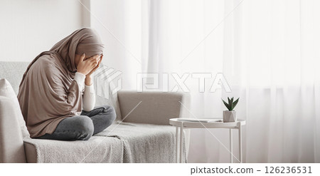 Depression concept. Upset muslim woman in hijab crying on sofa at home, hiding her face in hands, feeling desperate and lonely, free space 126236531