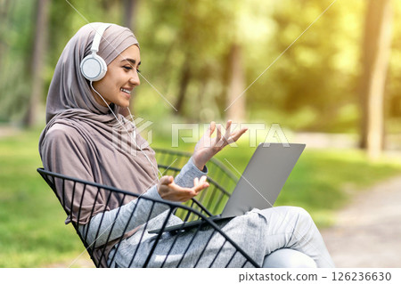 Pretty muslim girl having video conference while resting at park, using laptop and headset, empty space 126236630