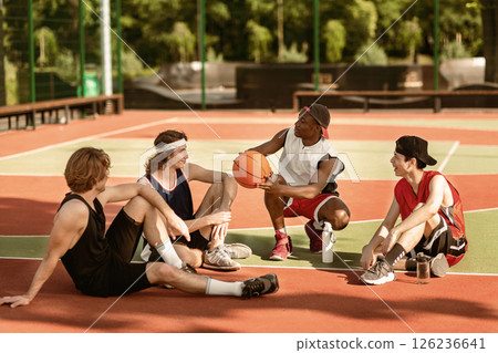 Team of young basketballers discussing their game strategy, sitting at outdoor stadium after training, free space. Group of millennial basketball players chatting to each other before friendly match Team of young basketballers discussing their game strategy, sitting at outdoor stadium after training, free space. Group of millennial basketball players chatting to each other before friendly match 126236641