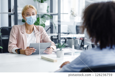 New normal and modern interview during covid-19 outbreak. HR manager with tablet looking for employee, watch at african american woman through protective glass in modern office interior, free space New normal and modern interview during covid-19 outbreak. HR manager with tablet looking for employee, watch at african american woman through protective glass in modern office interior, free space 126236759