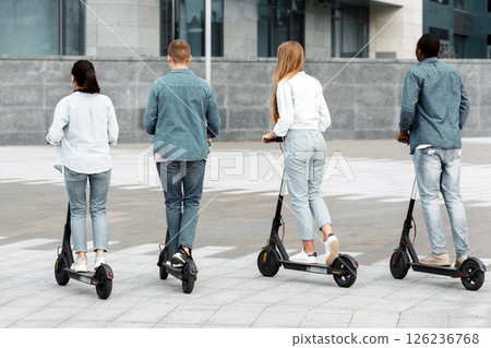 A group of diverse teenagers is riding scooters down a smooth urban street in broad daylight. The young riders are dressed casually and showcase a vibrant youth culture, back view A group of diverse teenagers is riding scooters down a smooth urban street in broad daylight. The young riders are dressed casually and showcase a vibrant youth culture, back view 126236768