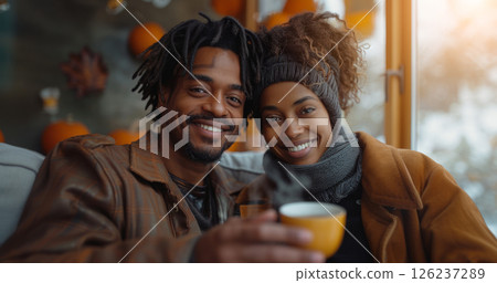 A couple smiles and looks at the camera while holding a cup of coffee. They are both wearing winter clothes and sitting by a window with a view of a snowy landscape. 126237289