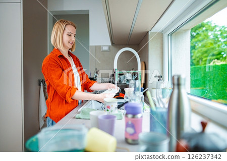 Woman happily cleaning dishes in a sunny kitchen Woman happily cleaning dishes in a sunny kitchen 126237342